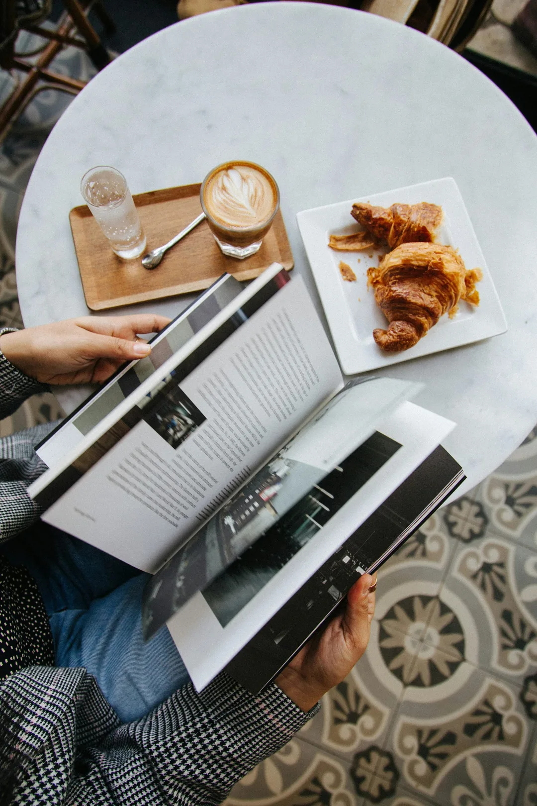 A person reading a magazine at a café table beside a coffee and a croissant