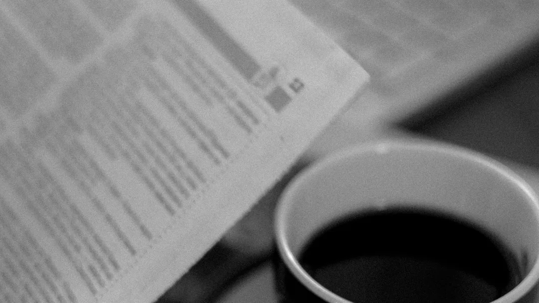A newspaper folded beside a cup of coffee, in black and white