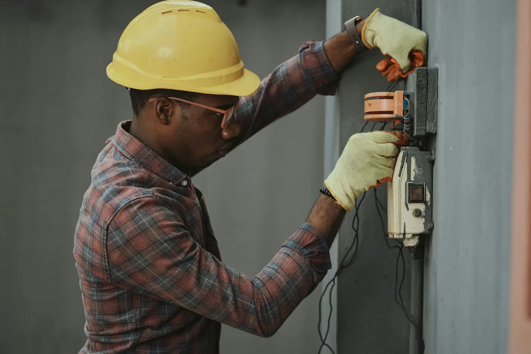 An electrician in a yellow hard hat and gloves working on a wall-mounted electrical panel