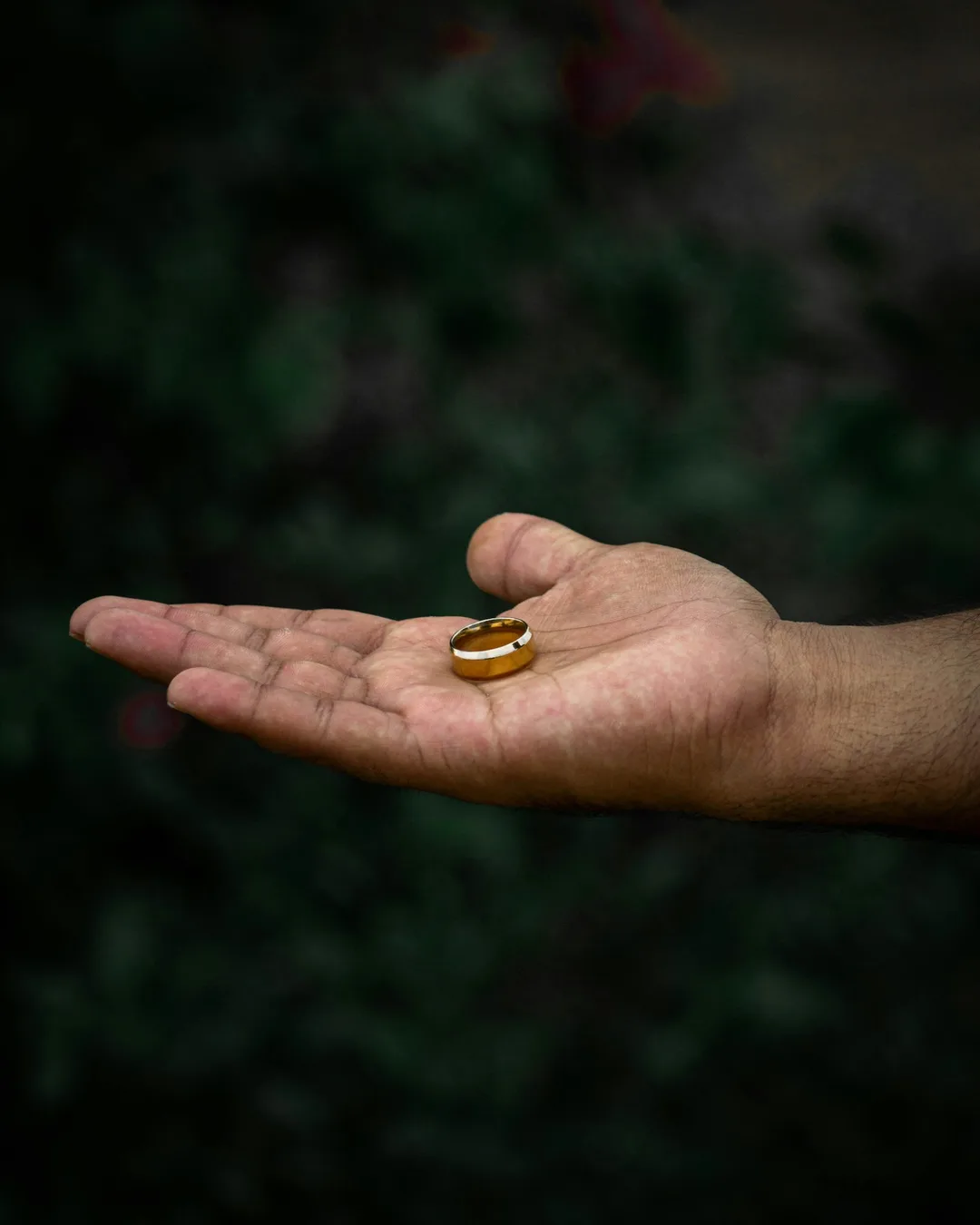 An open hand holding a plain gold band, against a dark green background